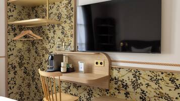 Une chambre d’hôtel avec un bureau en bois, une chaise, un téléviseur fixé au mur et un papier peint à motifs floraux.