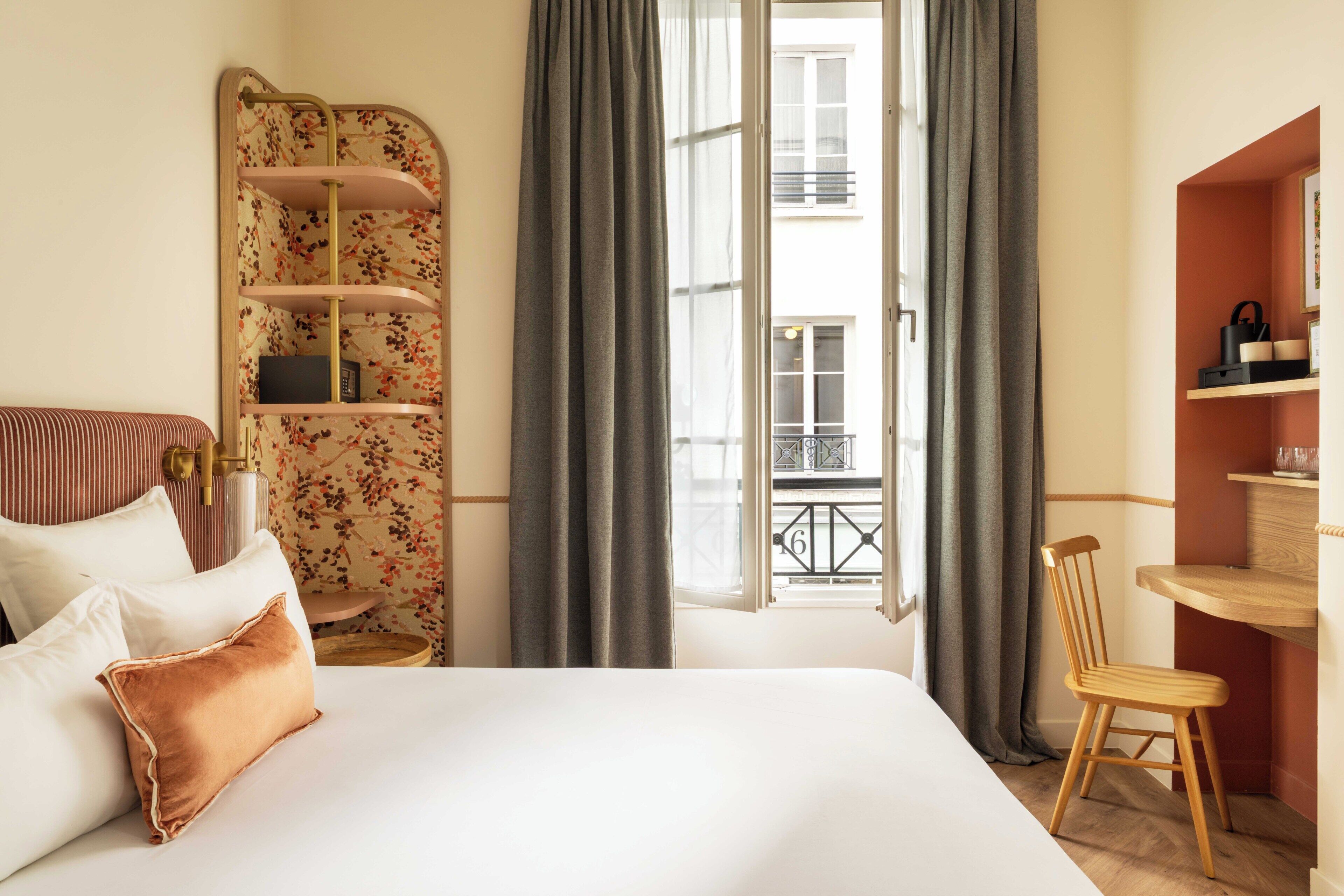 A hotel room with a bed, a wooden chair, a small table, a bookshelf, and a balcony view.