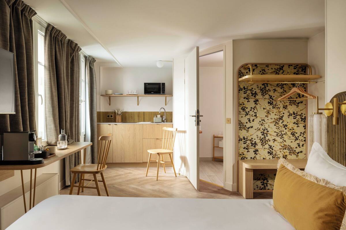 A hotel room with a bed, a desk, a chair, a kitchenette, and a bathroom.