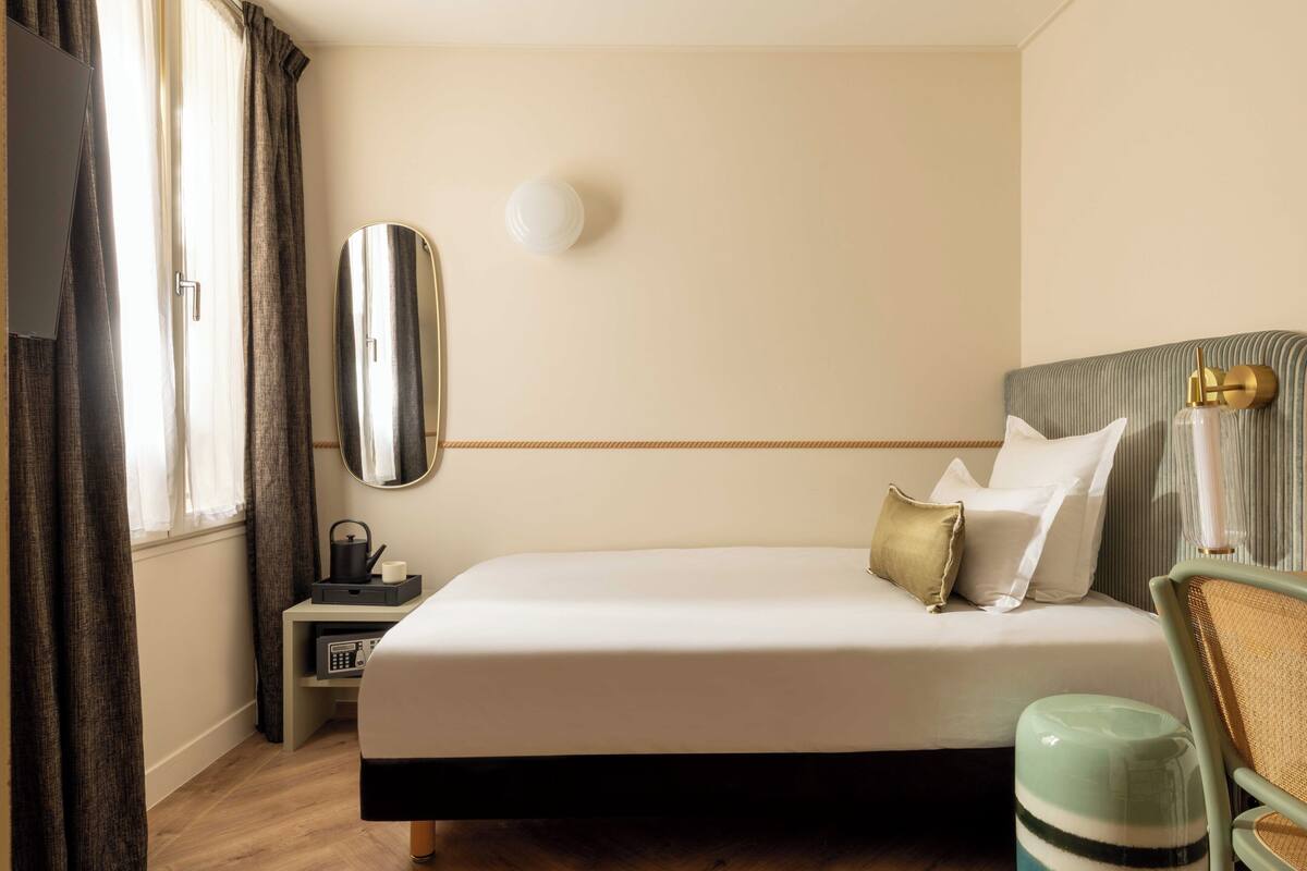 A hotel room with a bed, bedside table, lamp, and a view of the outdoors.