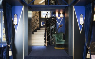 Un escalier menant à une pièce où se trouvent un fauteuil recouvert d’un tissu vert, une sculpture d’oiseau et des lampes fixées au mur.