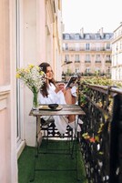 Une personne assise à une table, un livre à la main, sur un balcon d’où l’on aperçoit des bâtiments.