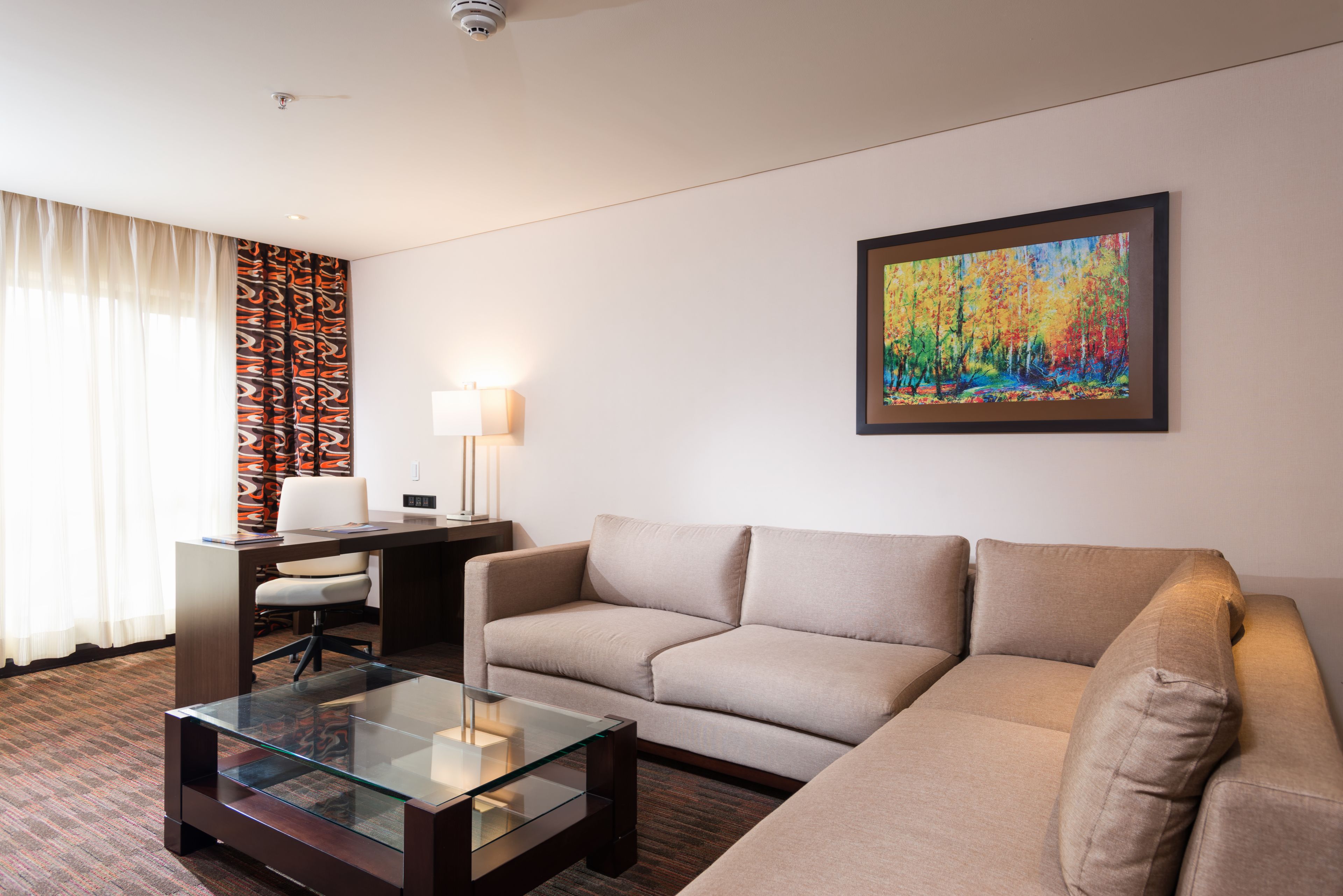 A modern hotel room with a sofa, a glass coffee table, a desk with a chair, a lamp, and a colorful painting on the wall.