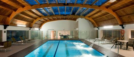 An indoor swimming pool with a glass ceiling, wooden beams, and lounge chairs.