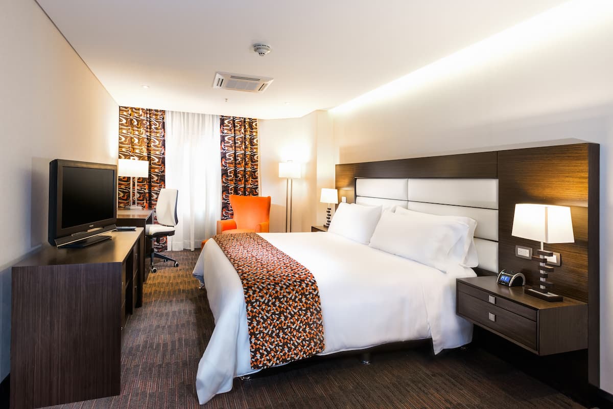 A hotel room with a bed, a desk with a TV, a chair, a nightstand, and a lamp.