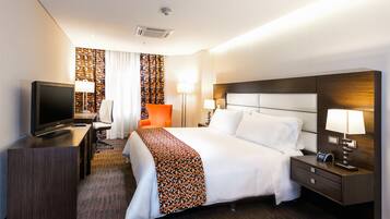 A hotel room with a bed, a desk with a TV, a chair, a nightstand, and a lamp.