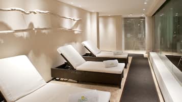 Moderni spa-huone, jossa on kolme lepotuolia, lasiseinällä varustettu suihku ja minimalistinen muotoilu.