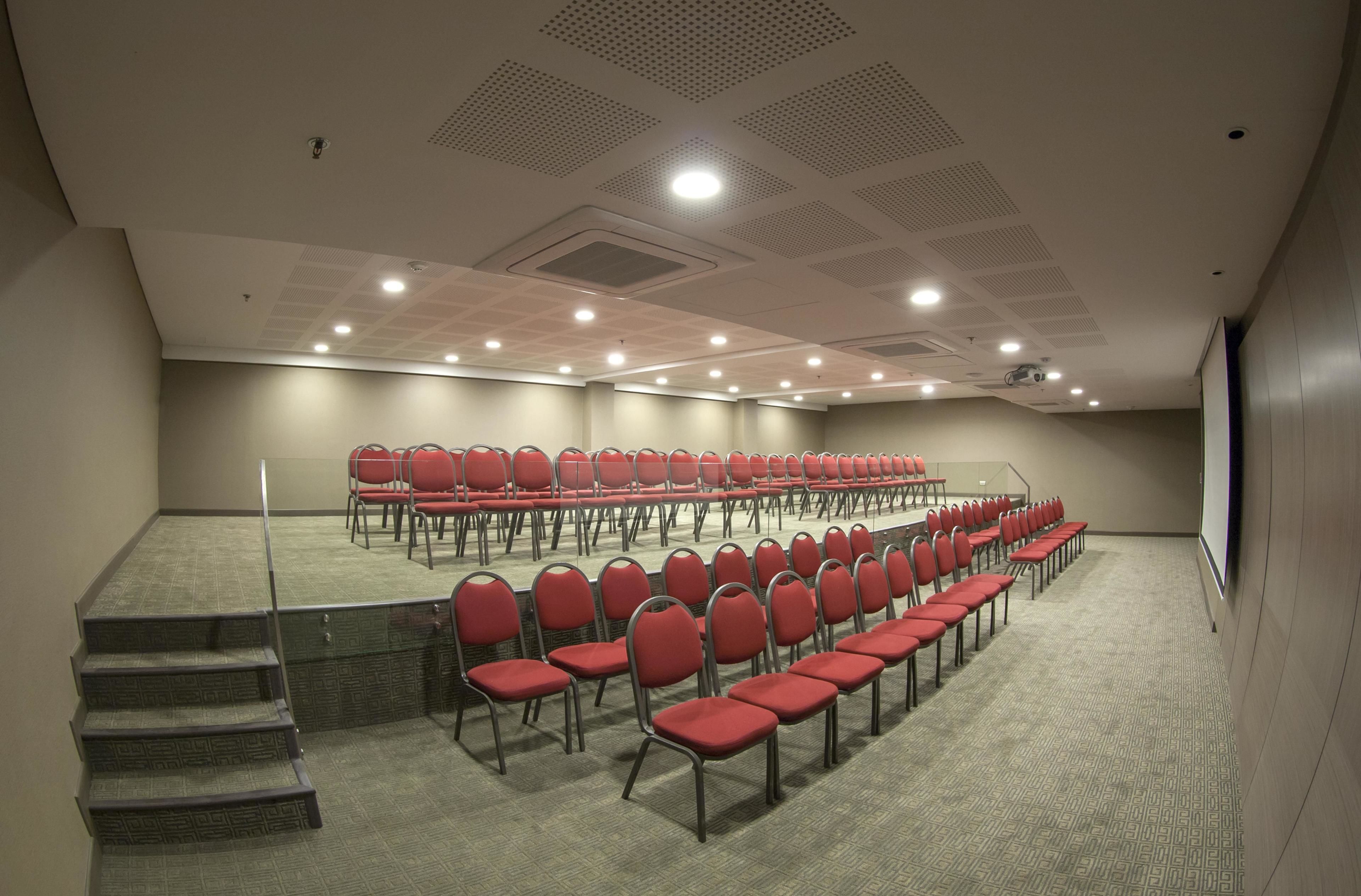 Une salle de conférence avec des rangées de chaises rouges, un sol recouvert de moquette et un écran de projection.