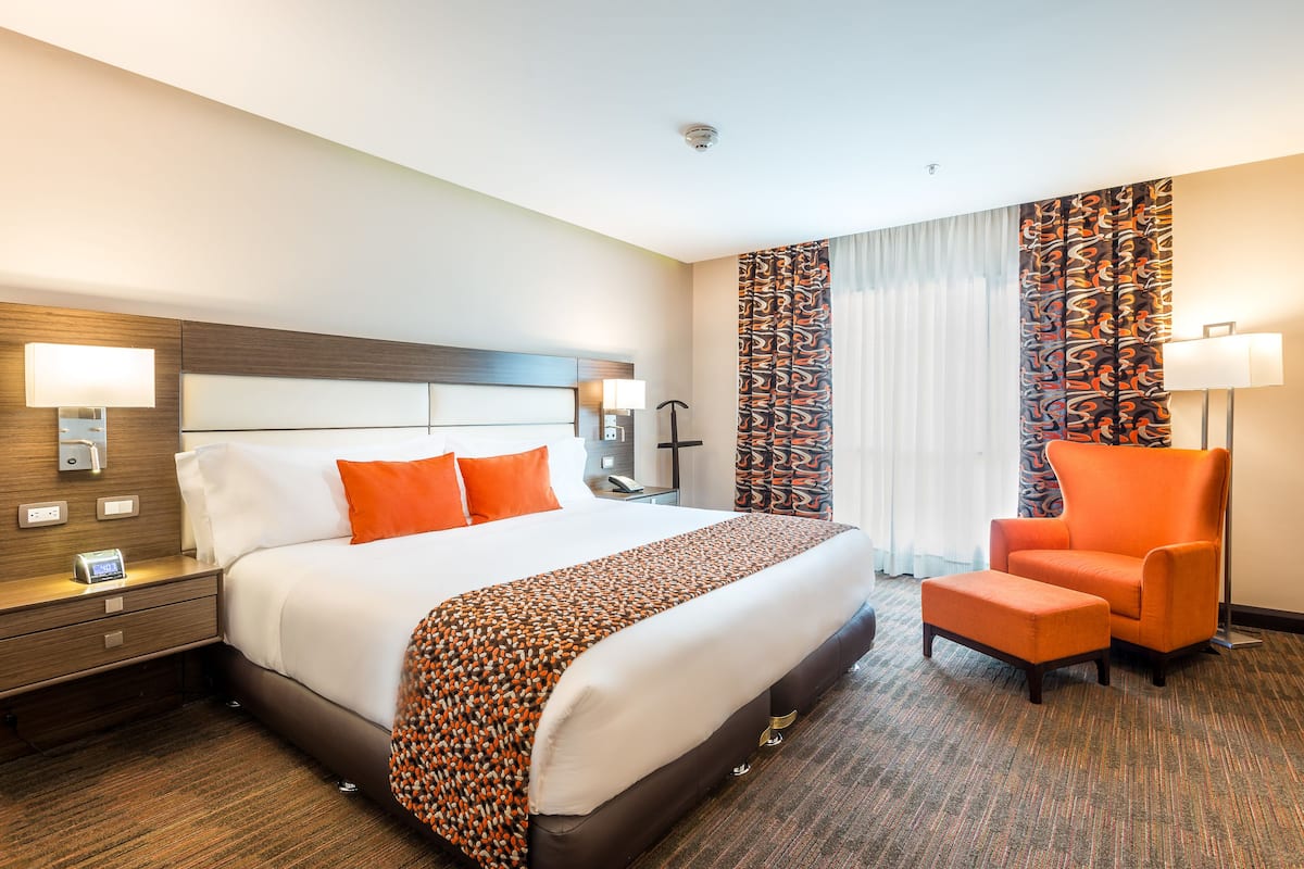 A hotel room with a large bed, an orange armchair, a nightstand with a lamp, and a patterned curtain.