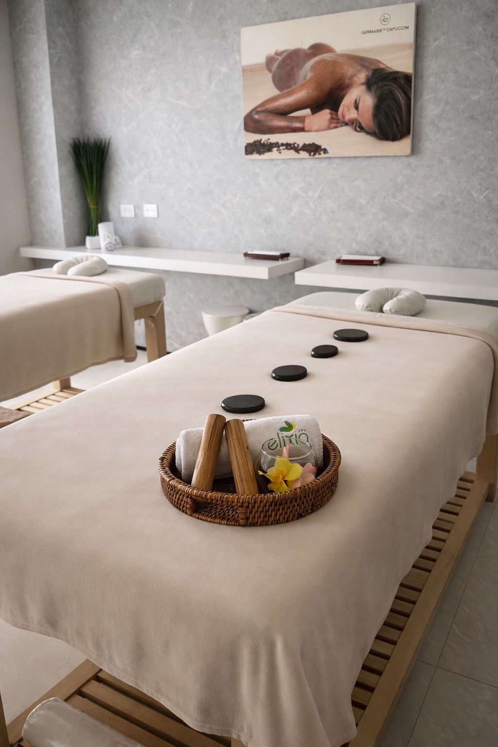 Un cuarto de spa con dos camillas de masaje, un cuadro en la pared y una cesta con toallas y un vaso.