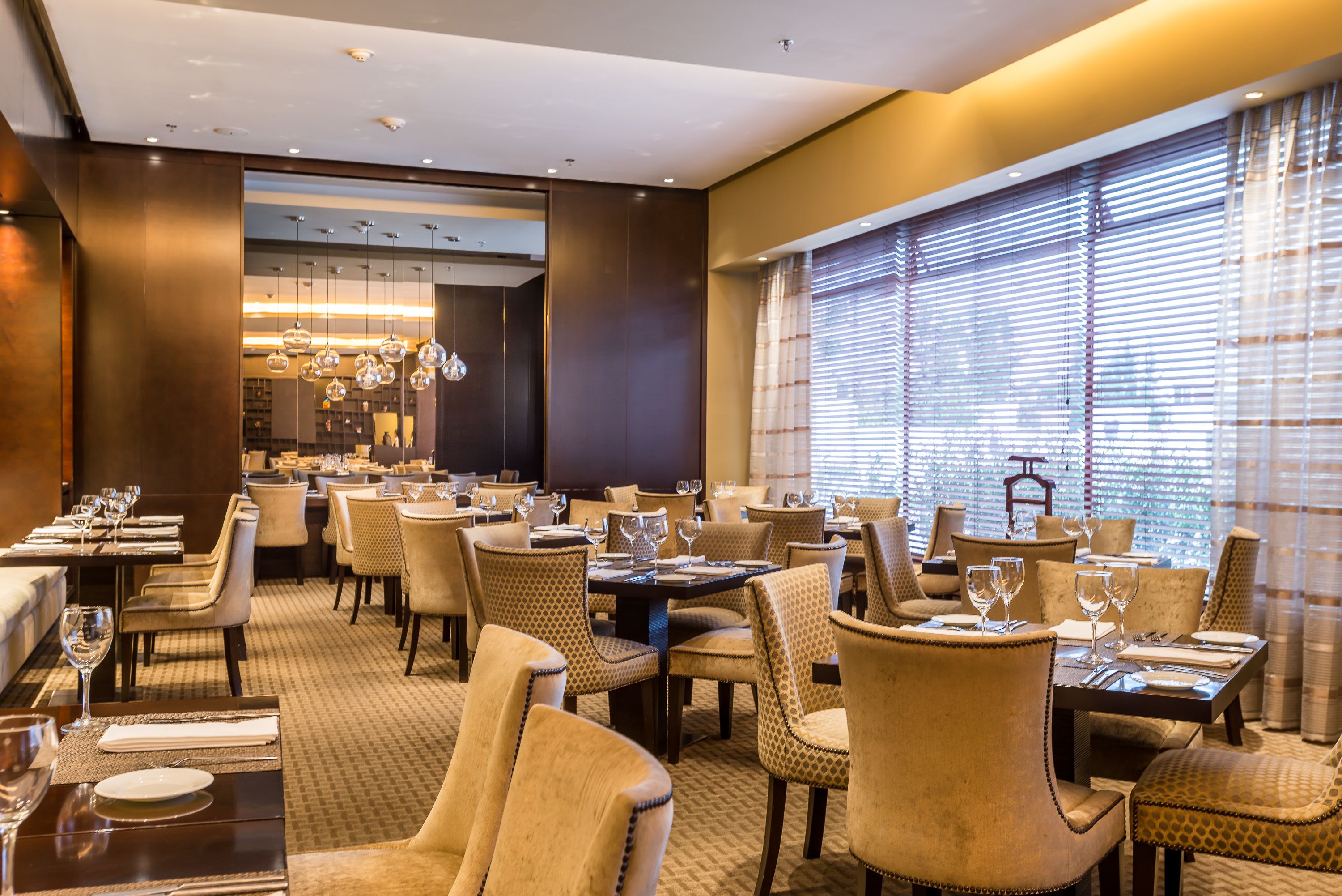 Une salle à manger d’hôtel avec de nombreuses tables et chaises, un lustre et de grandes fenêtres équipées de stores.