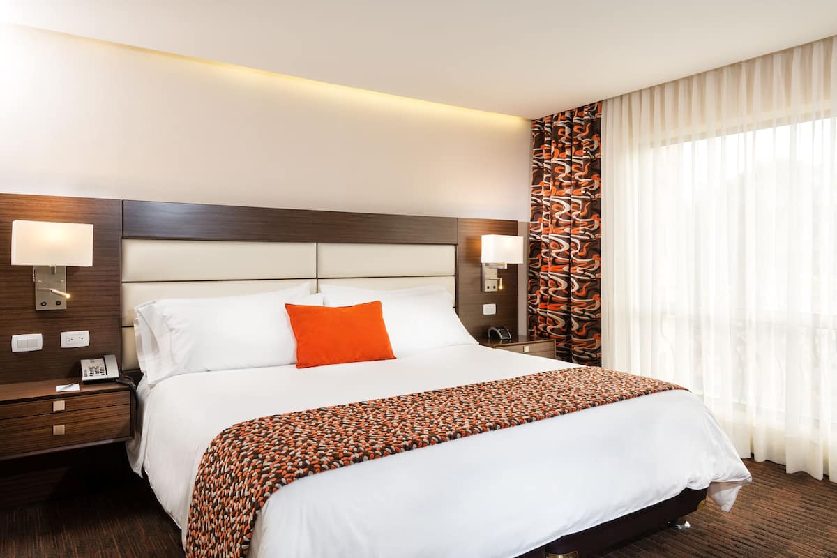 A hotel room with a large bed, two bedside tables, a nightstand, a patterned curtain, and a modern headboard.
