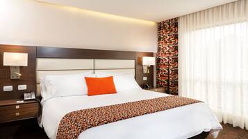 Habitación de hotel con una cama grande, dos mesitas de noche, una mesita auxiliar, una cortina estampada y un cabecero moderno.