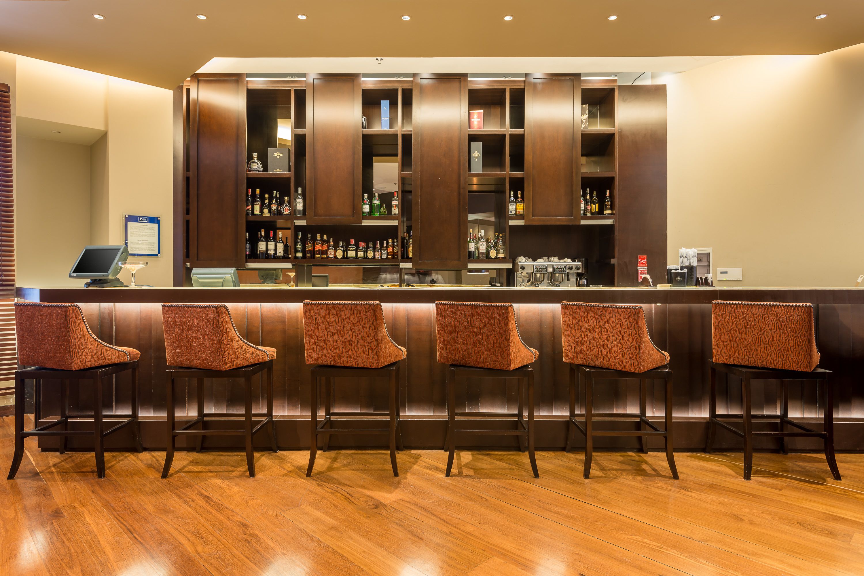 A bar area with a wooden counter, multiple shelves with bottles, and five upholstered bar stools.