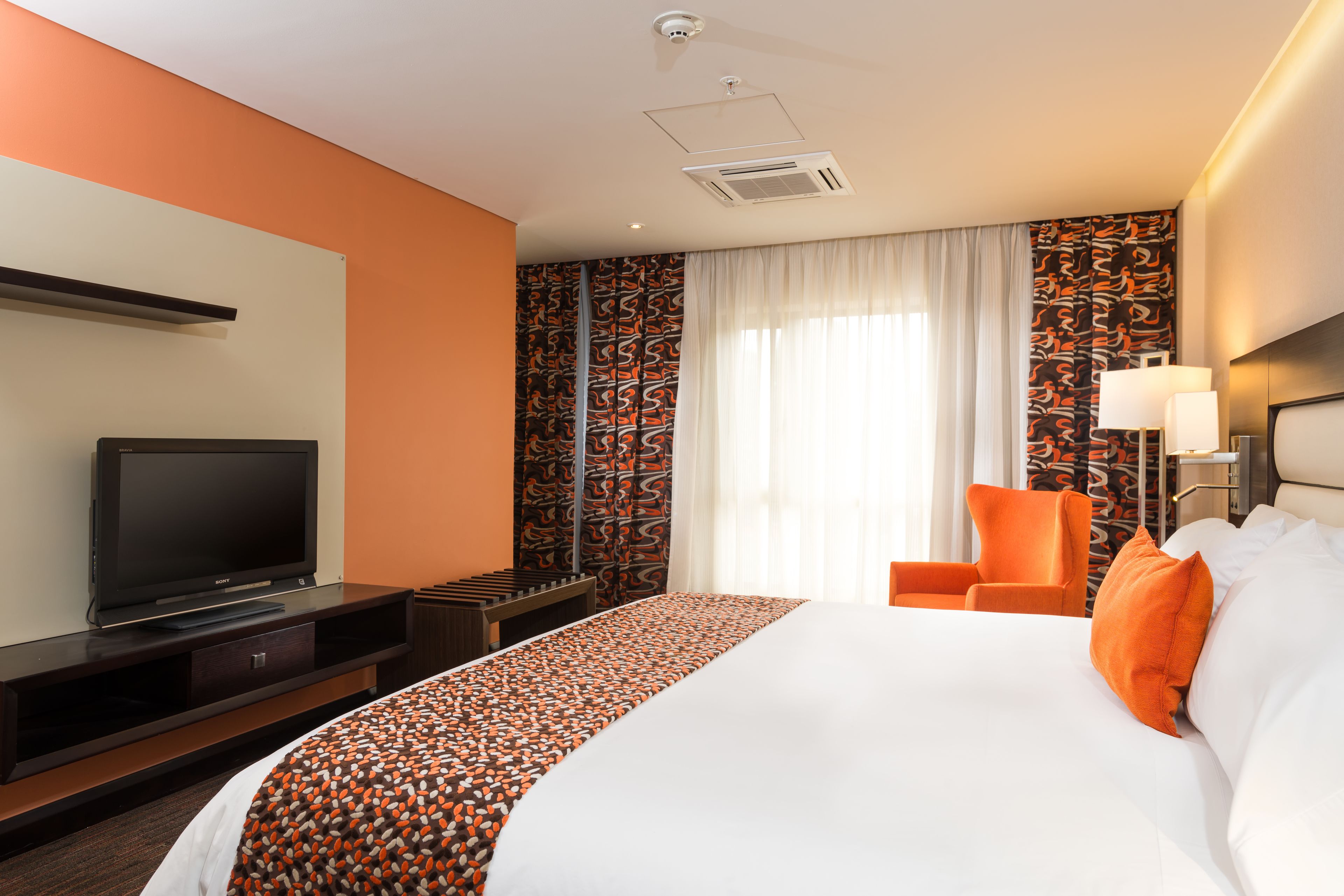 Habitación de hotel con una cama grande, un televisor sobre un mueble bajo, una silla naranja y cortinas estampadas.