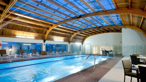 Una amplia piscina interior con techo de madera y paneles de vidrio, rodeada de sillones de descanso y un comedor.