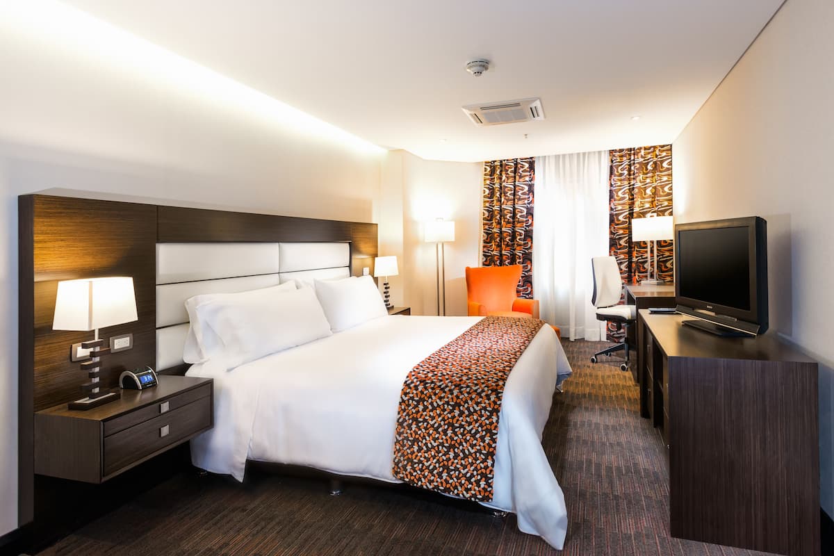 A hotel room with a large bed, a desk with a television, a nightstand with a lamp, and a patterned curtain.