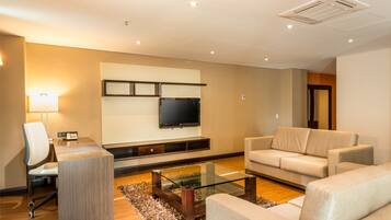 A modern living room with a flat-screen TV, a glass coffee table, a sofa, a desk with a chair, and a lamp.
