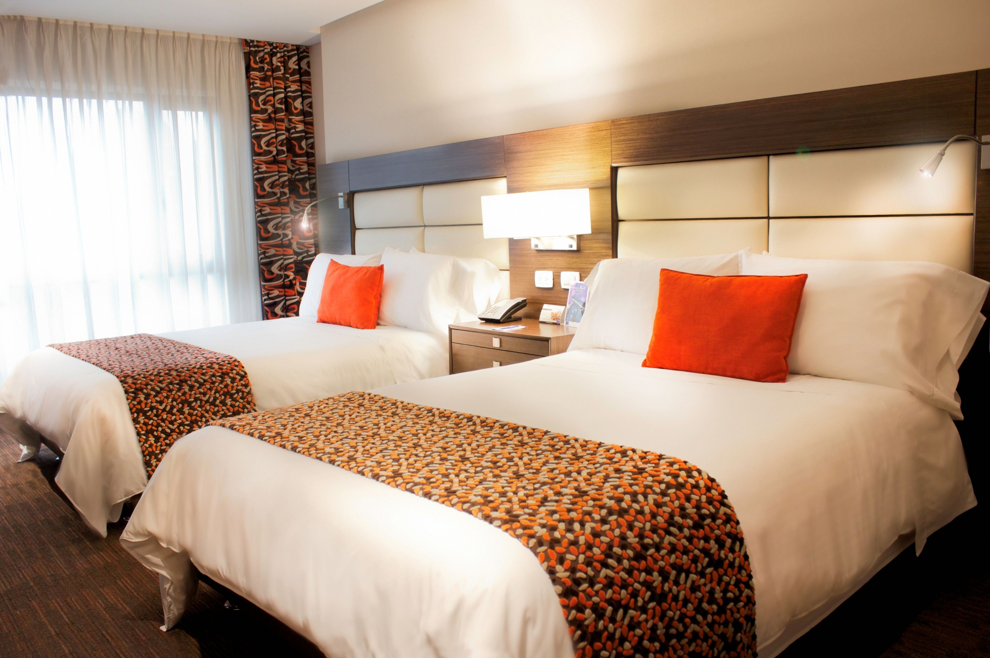A hotel room with two beds, each with white linens and orange accents. There is a nightstand with a lamp and a phone between the beds. The room has a wooden headboard and a window with curtains.