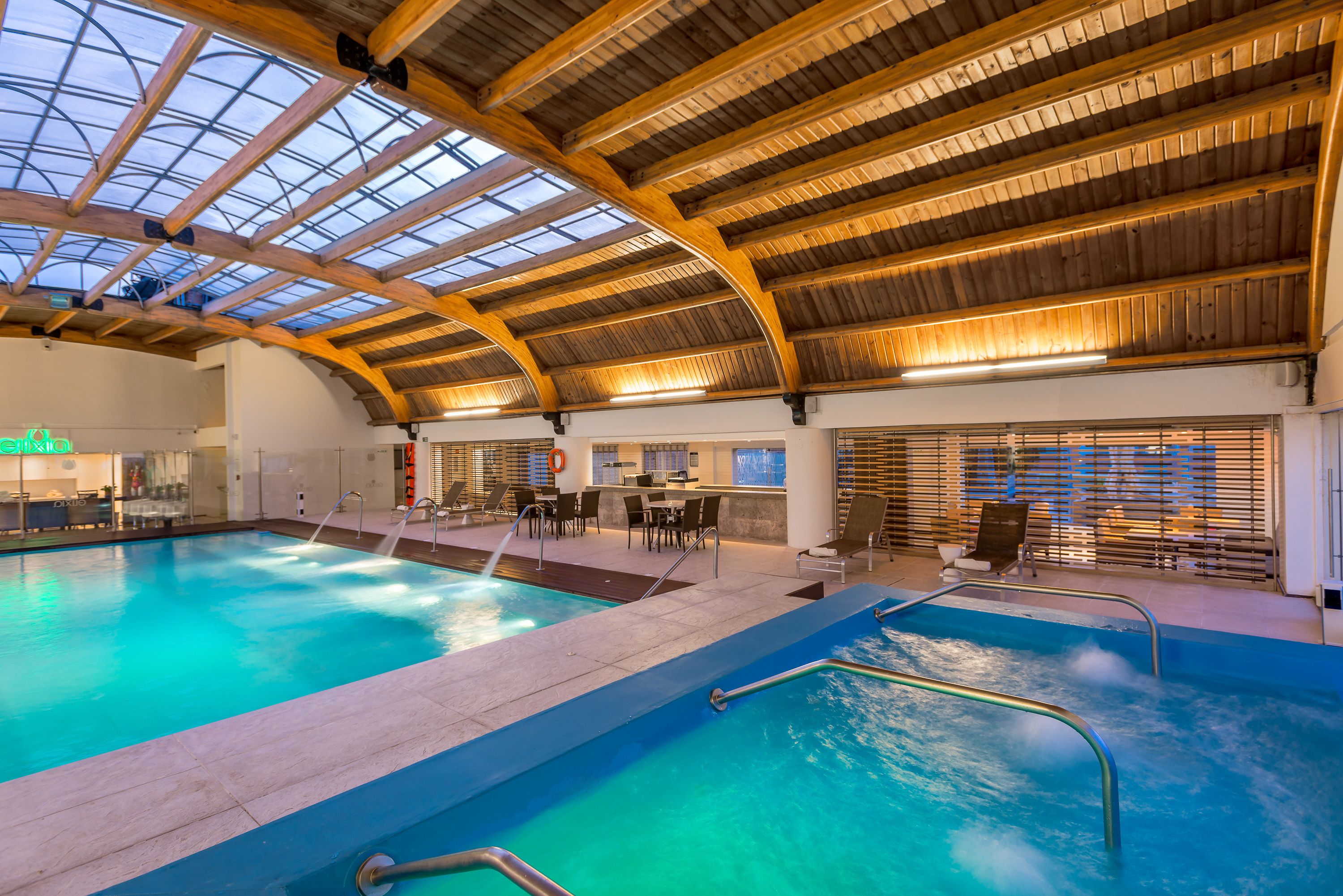 An indoor swimming pool with a glass ceiling, wooden beams, and a bar area with seating.