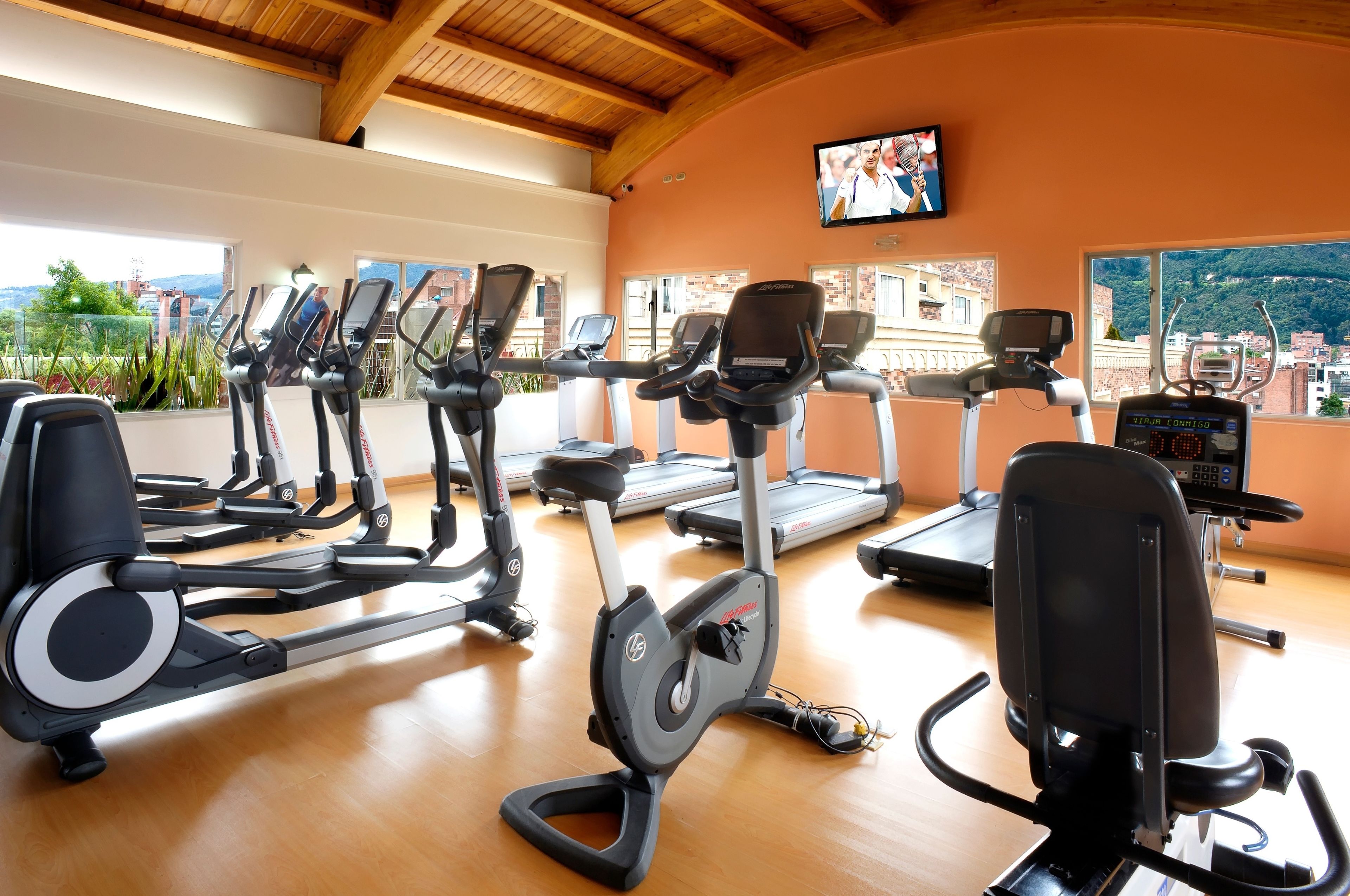 A gym with multiple exercise machines, large windows with a city view, and a television mounted on the wall.