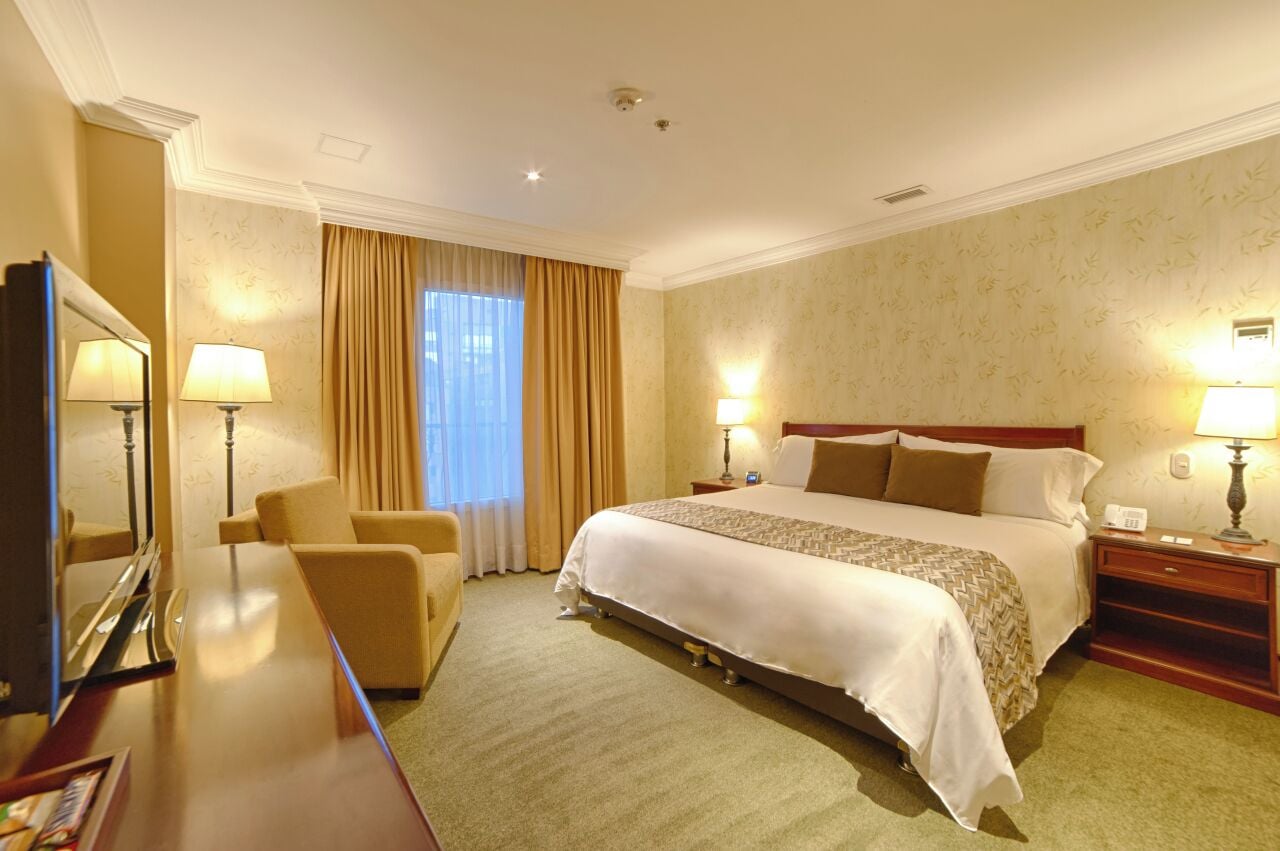 A hotel room with a large bed, two bedside tables with lamps, a chair, a desk with a mirror, and a window with curtains.