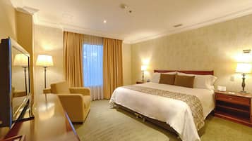 A hotel room with a large bed, two bedside tables with lamps, a chair, a desk with a mirror, and a window with curtains.