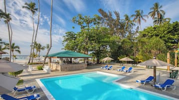 Ein Swimmingpool mit Liegestühlen und Sonnenschirmen, umgeben von Palmen und Blick auf einen Strand.