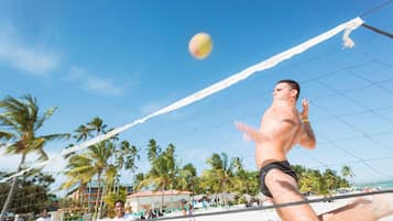 Un match de volleyball de plage avec des joueurs et des spectateurs, des palmiers et un ciel dégagé.