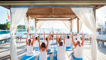 Eine Gruppe von Menschen praktiziert Yoga auf blauen Matten unter einem hölzernen Pavillon mit weißen Vorhängen, mit Blick auf ein Gewässer.