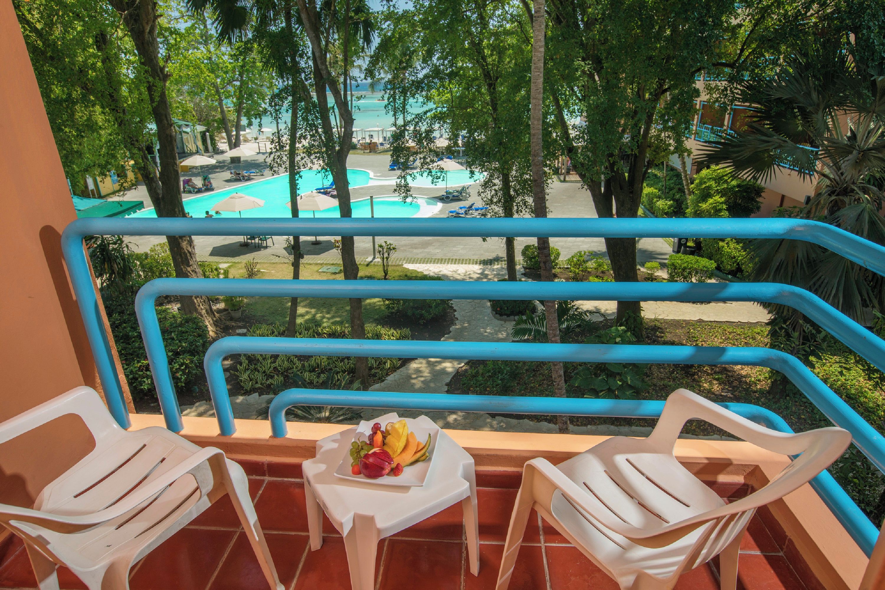 A balcony with two white plastic chairs, a small table with fruit, and a view of a swimming pool and resort buildings.