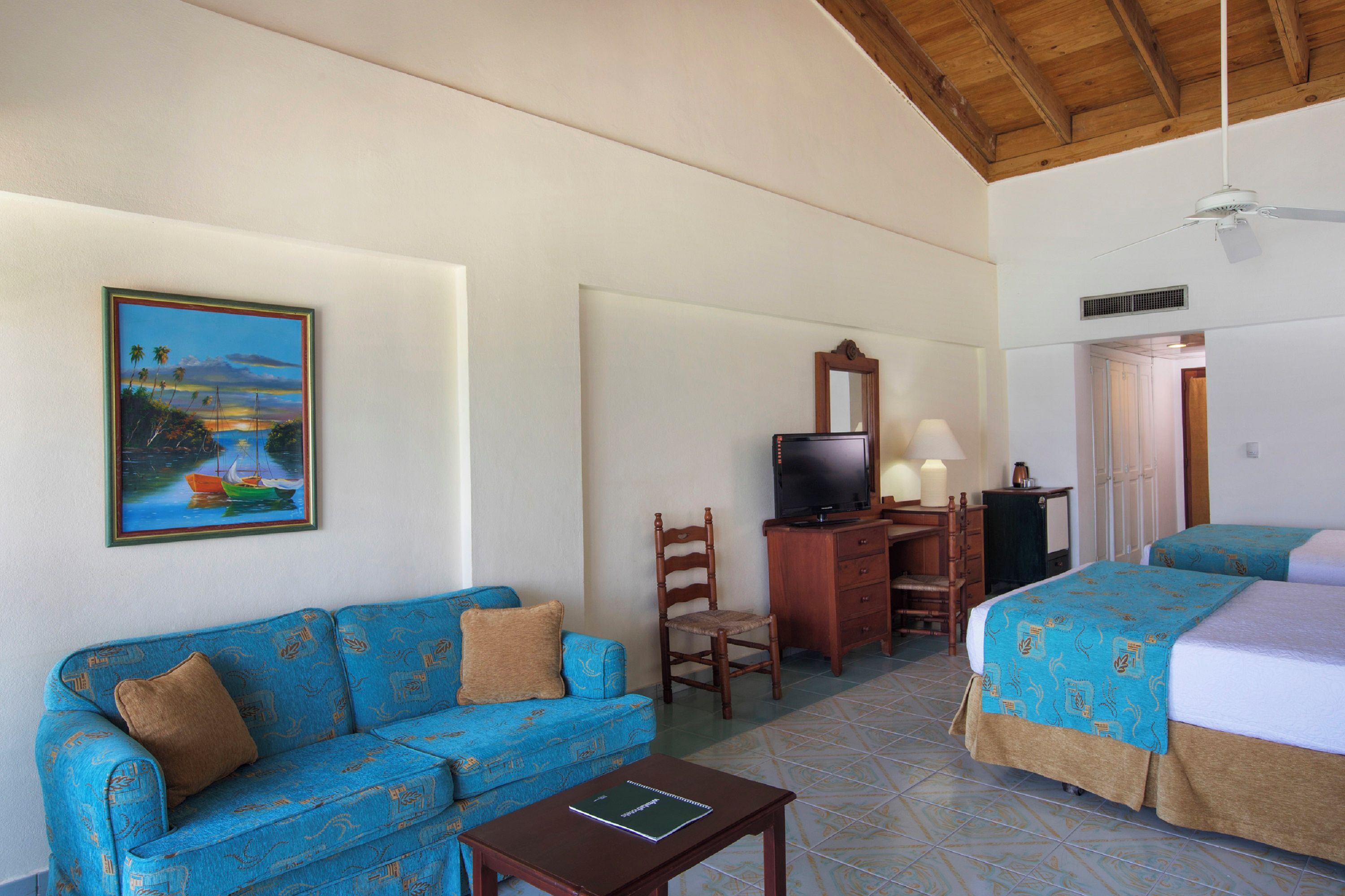 Una camera d'albergo con un divano blu, una scrivania con televisione, un letto con copripiumino decorato e un quadro raffigurante una costa.
