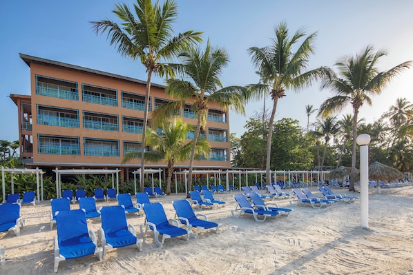 A hotel with multiple floors, a beach area with blue lounge chairs, and palm trees.