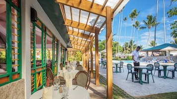 Un espace extérieur aménagé pour les repas, avec des tables et des chaises, une piscine en arrière-plan et des palmiers.