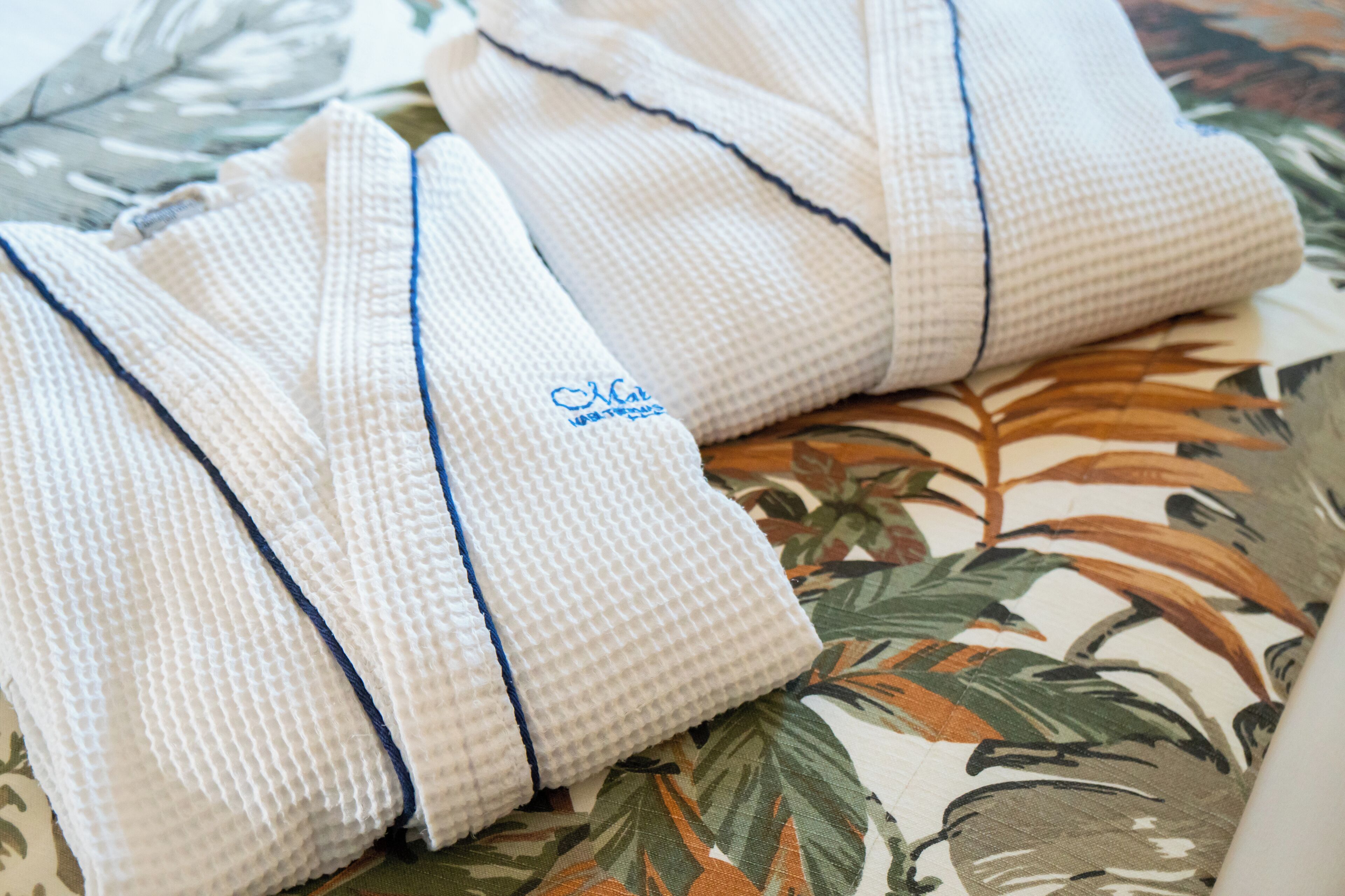 Una toalla blanca doblada con detalle, con un borde azul y un logotipo sobre un edredón estampado con motivos florales.