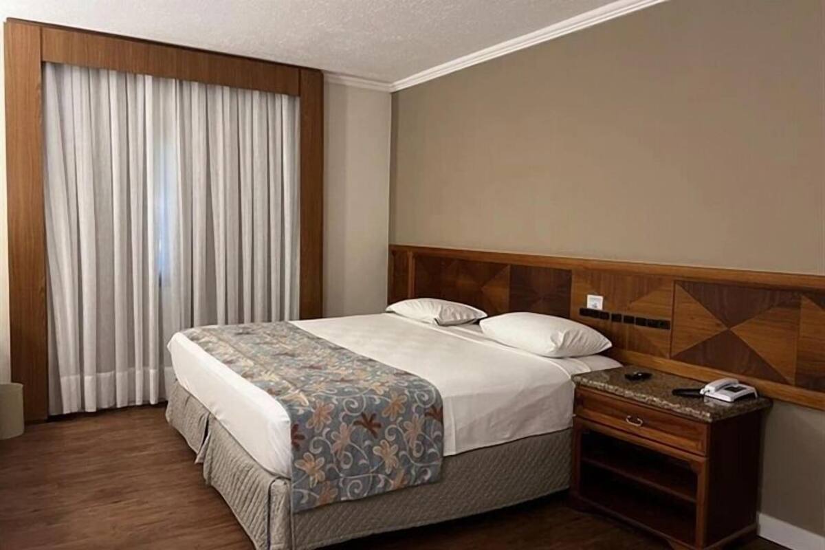 A hotel room with a bed, a nightstand, a wooden headboard, and a window with curtains.
