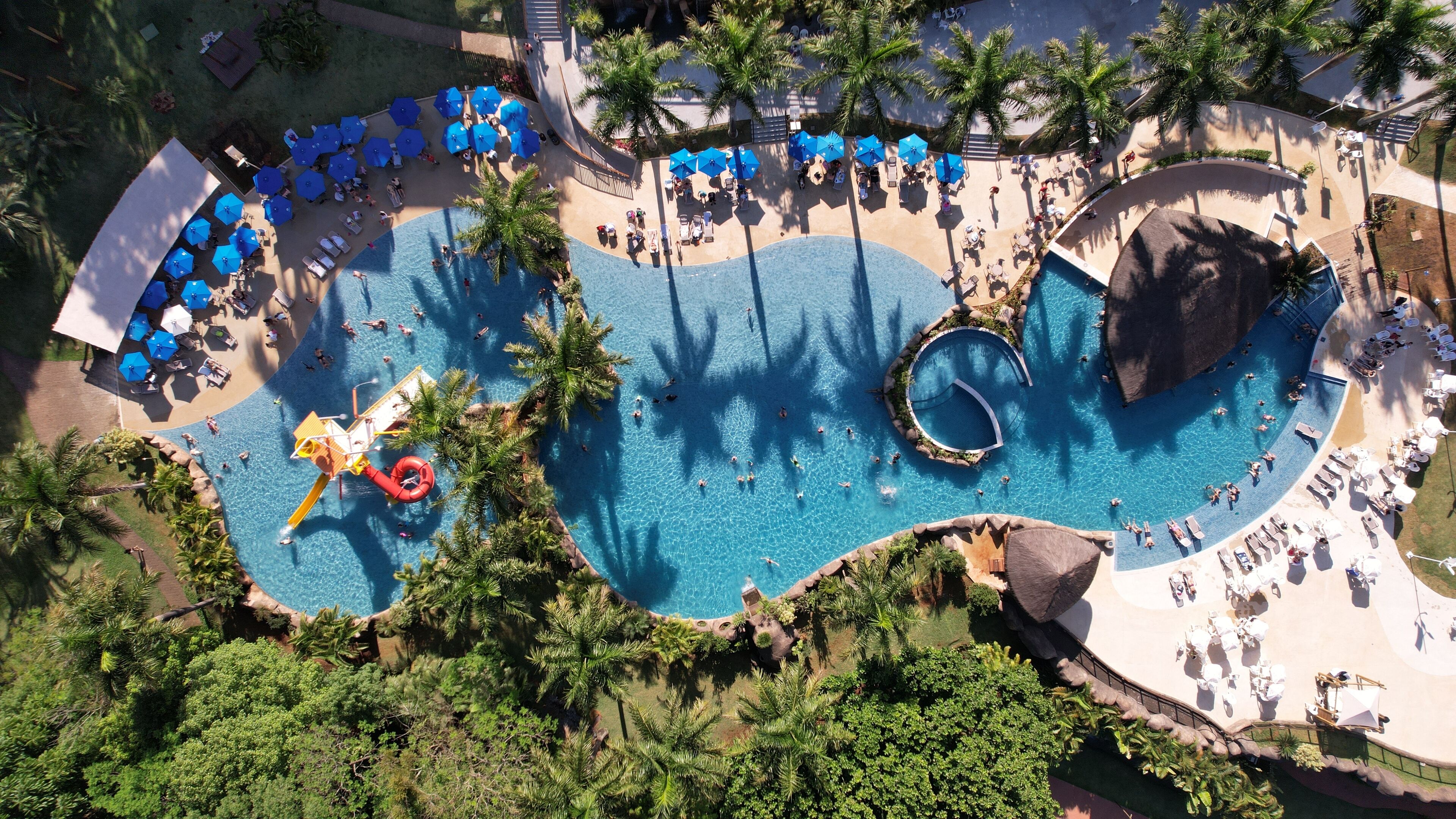 An aerial view of a resort with a large pool area, multiple lounge areas with umbrellas, and a water slide.