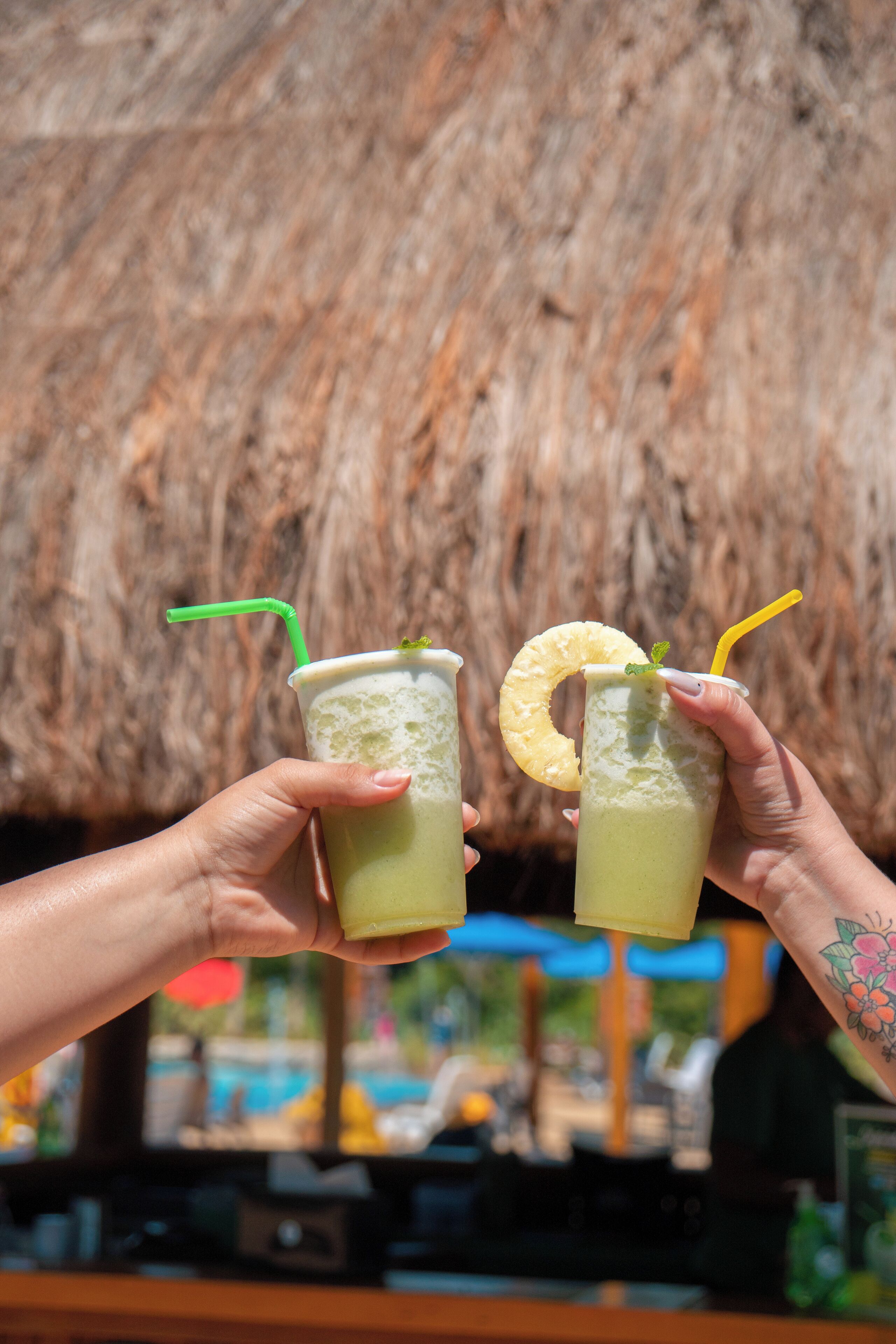 Two hands holding iced drinks with straws and pineapple slices.