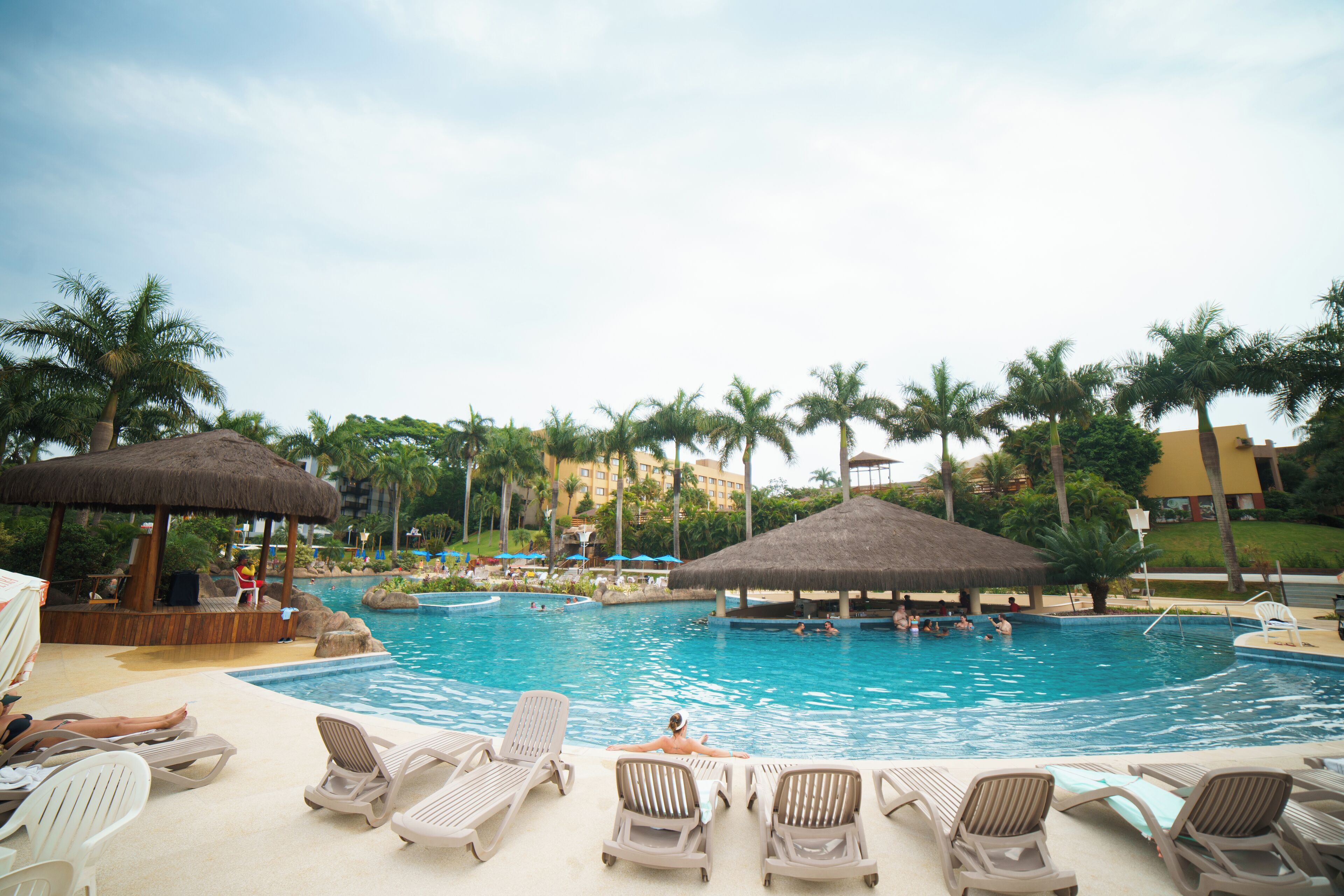 Zona de piscina con sillones, palmeras y sombrillas de paja.