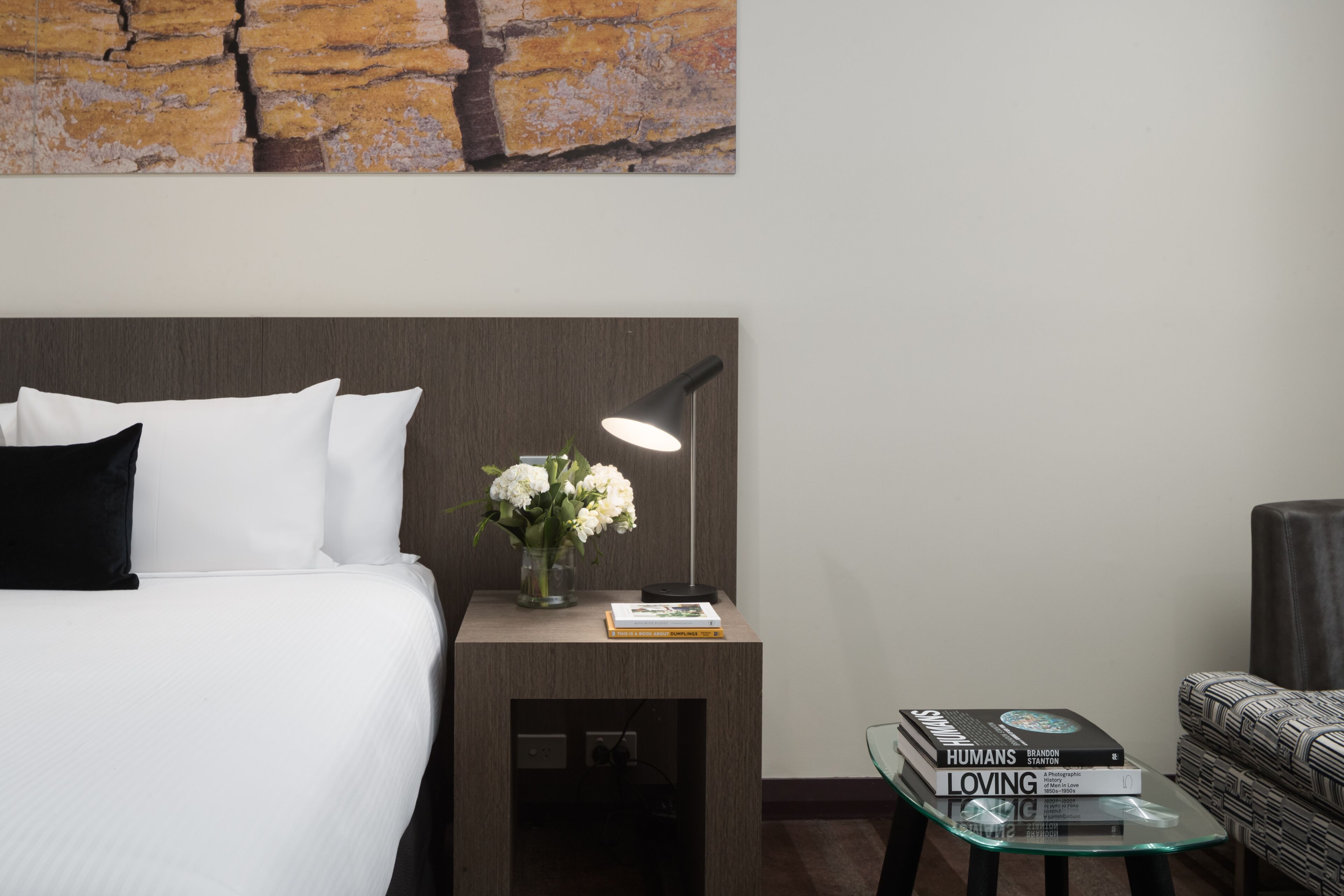 A neatly arranged hotel room with a bed, bedside table, lamp, and a small glass table with books.