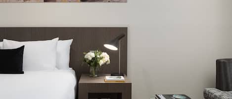 A neatly arranged hotel room with a bed, bedside table, lamp, and a small glass table with books.