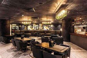 Un restaurante moderno con mesas de madera y sillas negras, una zona de bar con estantes llenos de botellas y una lámpara decorativa.