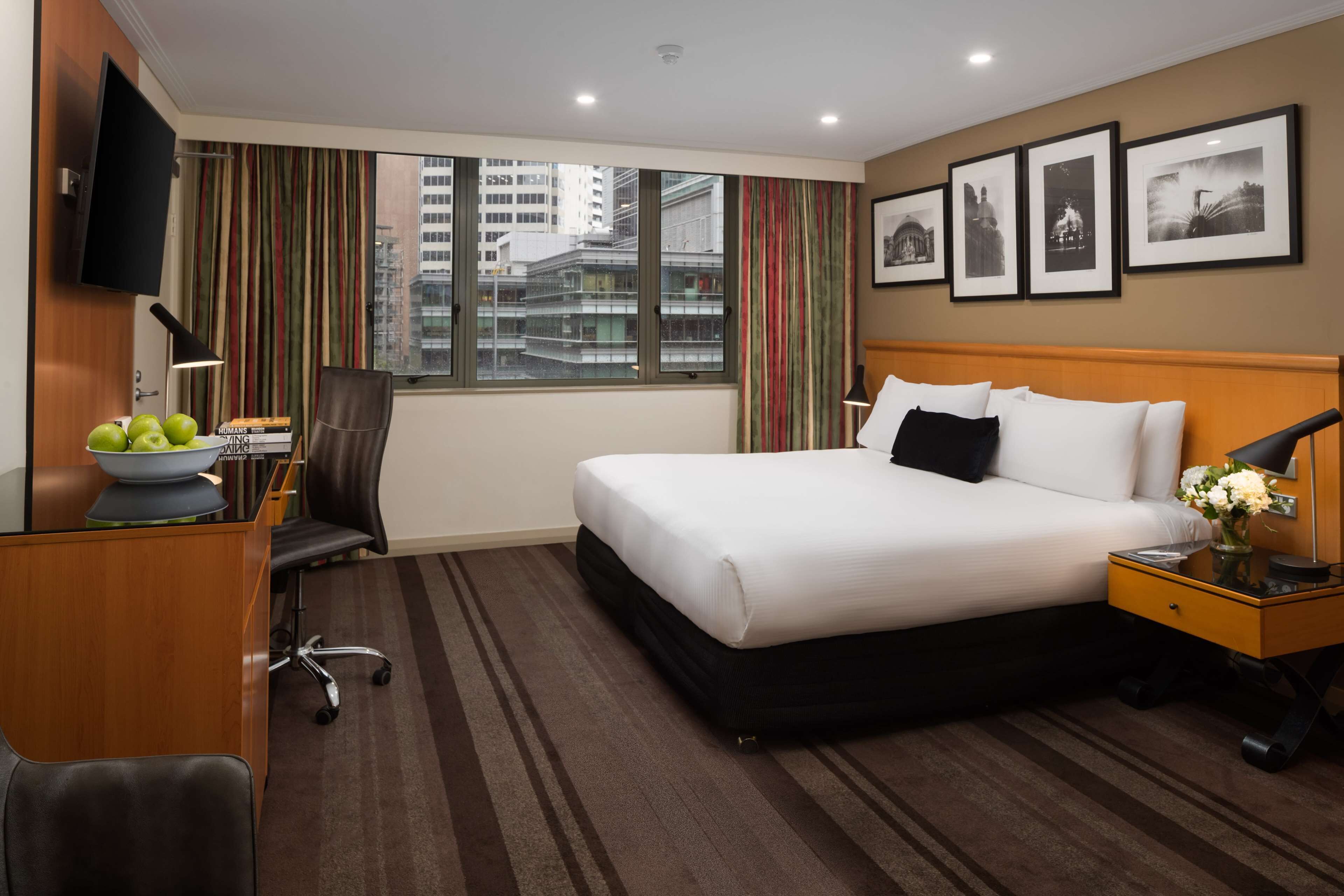 A hotel room with a large bed, a desk with a computer, a chair, a TV mounted on the wall, and a window with curtains.