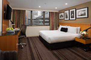 A hotel room with a large bed, a desk with a computer, a chair, a TV mounted on the wall, and a window with curtains.