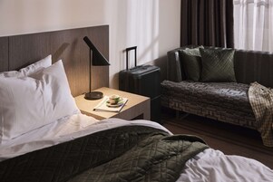 Una habitación de hotel con cama, mesita de noche con lámpara, maleta y sofá con manta.