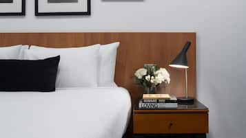 Ein ordentlich bezogenes Bett mit weißen und schwarzen Kissen, ein Nachttisch mit einer schwarzen Lampe, eine Vase mit weißen Blumen und ein Stapel Bücher mit dem Titel „HUMANS LOVING“.