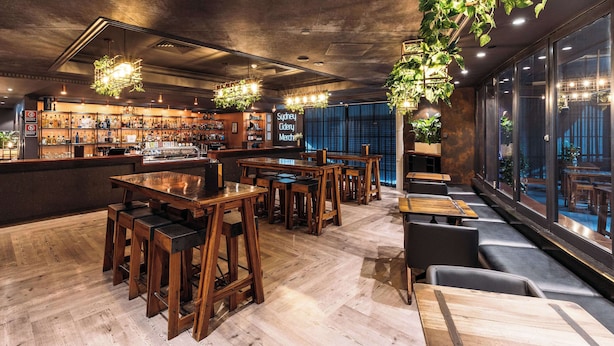 Een bar met houten meubels, hangplanten en een toog met verschillende flessen.