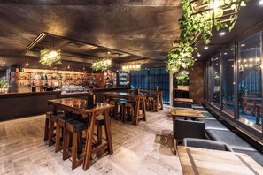 Een bar met houten meubels, hangplanten en een toog met verschillende flessen.