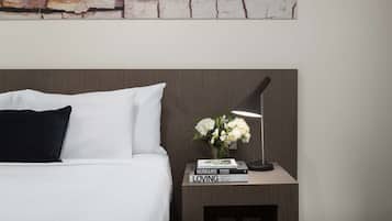 A neatly made bed with white and black pillows, a small bedside table with a lamp, a vase with white flowers, and a book titled "HUMANS LOVING" by Brandon Stanton.