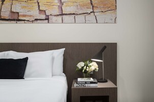 A neatly made bed with white and black pillows, a small bedside table with a lamp, a vase with white flowers, and a book titled "HUMANS LOVING" by Brandon Stanton.